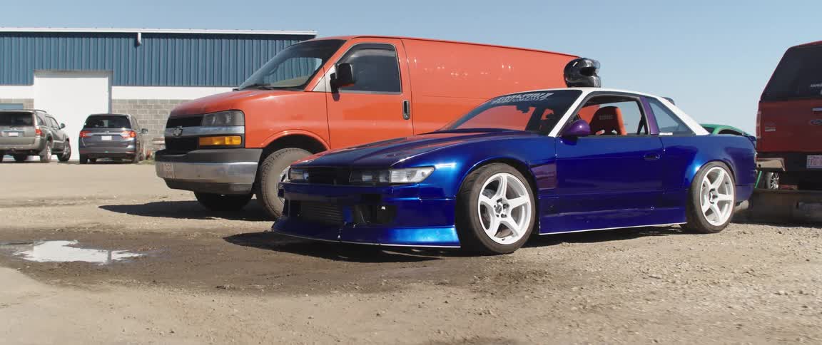 A blue sports car with a lowered suspension and aftermarket wheels is parked in a lot