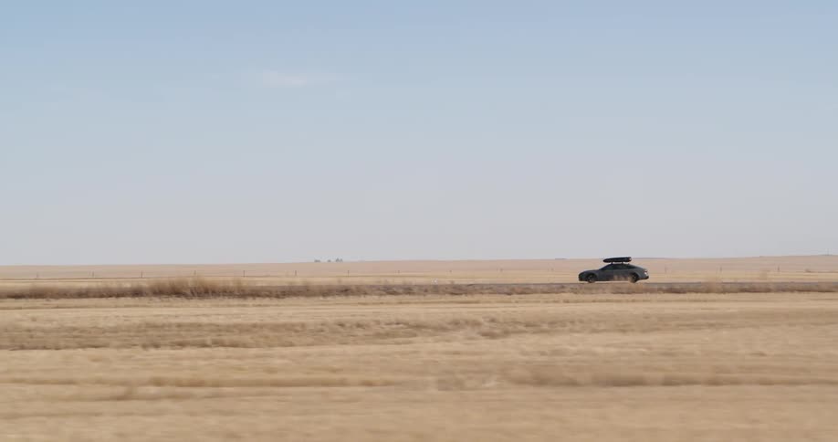 A car with a suitcase of luggage on top driving on a street in a desert region with dry grass
