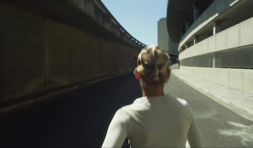 The back of a woman who is running on an asphalt path between an overpass and a building