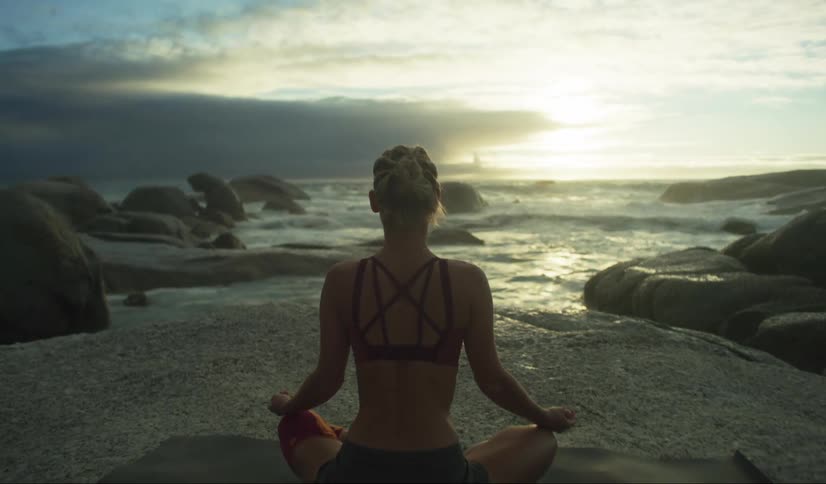 A woman is sitting crosslegged on a rocky beach facing the ocean during sunset