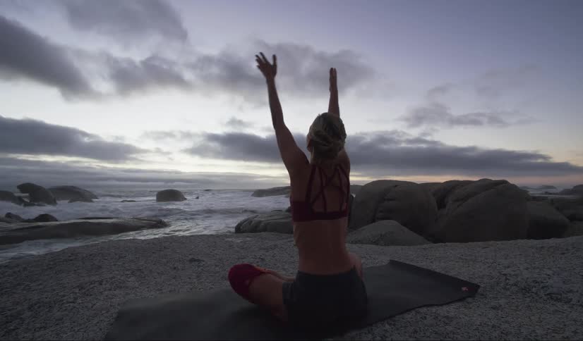 A woman is shown from behind sitting on a yoga mat stretching on the beach