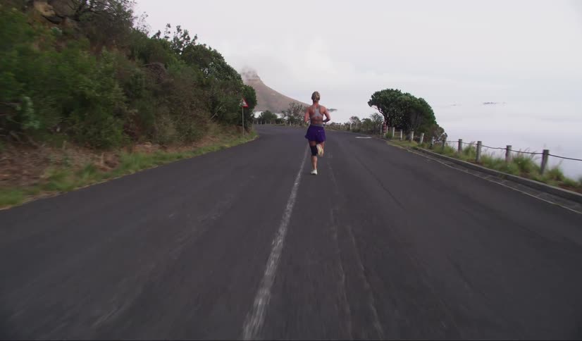 Blonde woman running on a paved road with mountains in the background and a cloudy sky