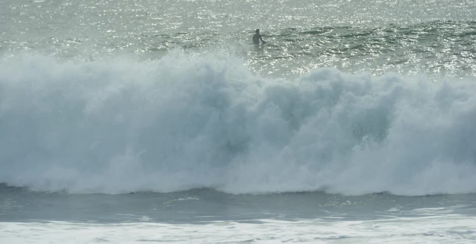 A big wave rolls in and two people appear on the water in the background