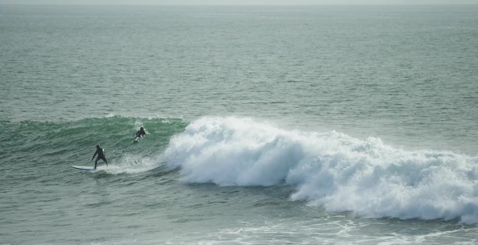A surfer rides a wave while another paddles over it