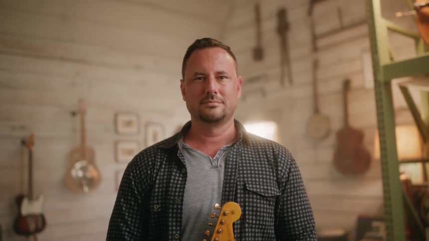 Man smiles slightly while standing with a guitar in a music room