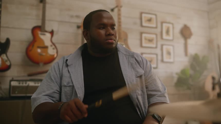 A bearded drummer in a black top plays drums in a room with guitars on the wall