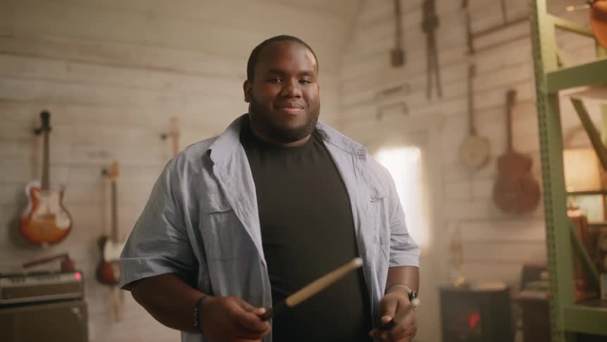 Bearded man wearing a black top and a blue shirt smiles while holding drumsticks