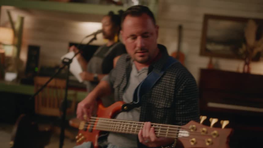 A cozy intimate music session is taking place in a warmly lit rustic room with a drummer, bassist, guitarist and singer performing together
