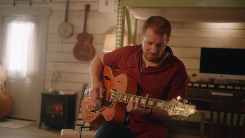 A bearded musician wearing a red shirt strums an acoustic guitar