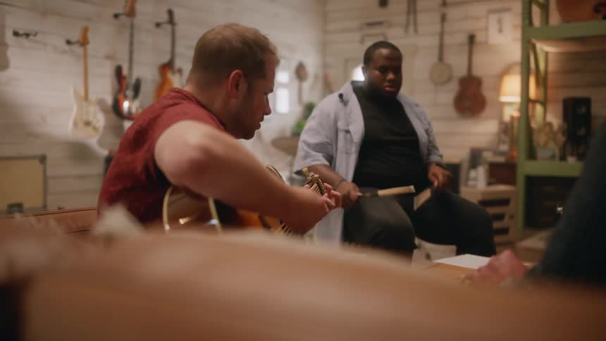 A bearded musician wearing a red shirt strumming a guitar next to a drummer playing drums in a professional music studio