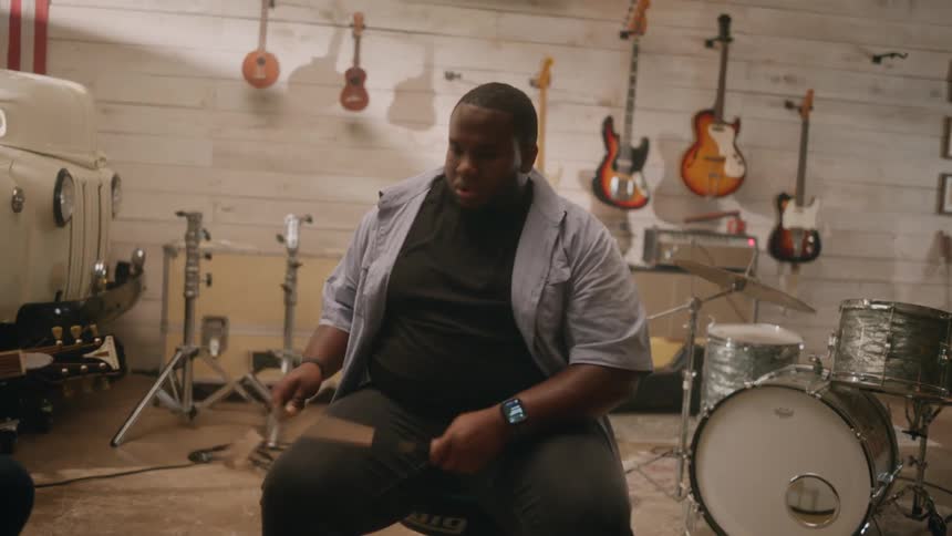 A man sits on a chair practicing on his knee with drumsticks in hand
