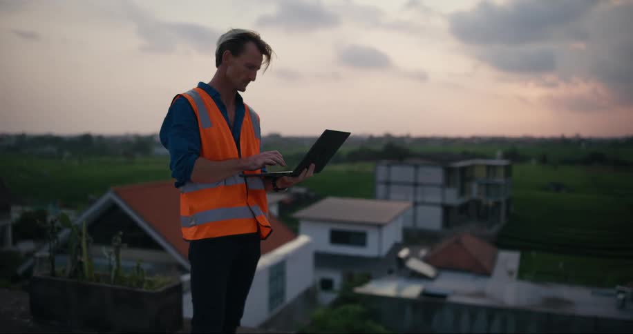 Engineer observes his surroundings outdoors and enters information on his laptop computer