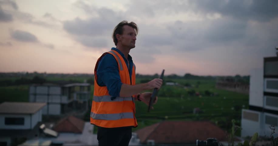 A worker in an orange and safety vest stands on a rooftop using a laptop