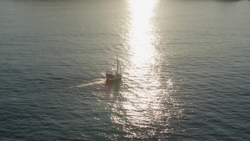 A small sailboat with white sails glides across a calm ocean at sunset