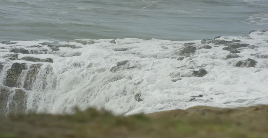 Waves crash then recede over a rocky outcropping during the day