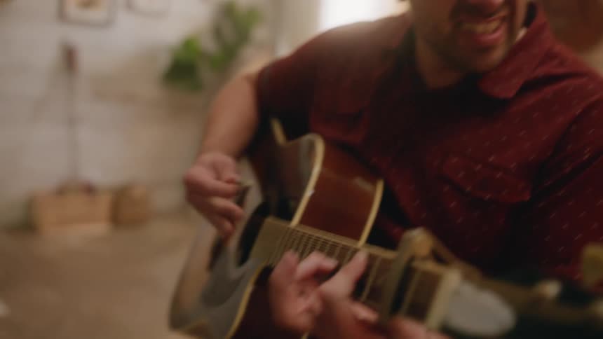 A bearded man in a red shirt plays a guitar while singing a song