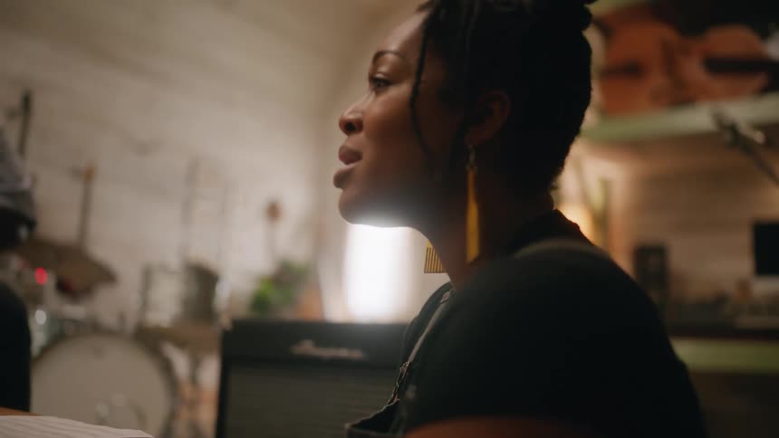 A woman with dreadlocks and yellow earrings sings a song around a table with the other musicians