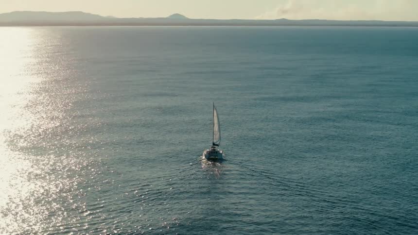 Sailboat sails toward the wide strip of water reflecting the bright sunshine