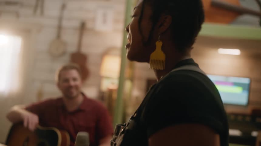 Woman holding a microphone and happily talking to a colleague next to her who is sitting with the guitar