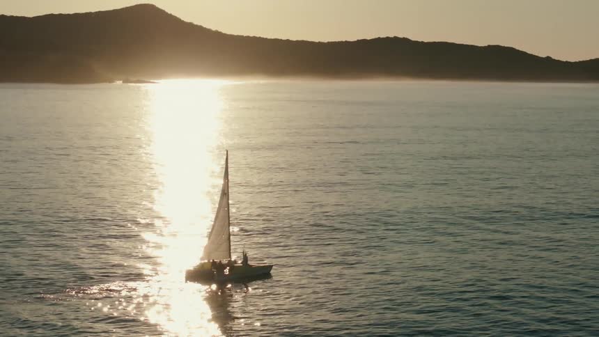 Sailing boat in the middle of the ocean with the coast and sunset on the horizon