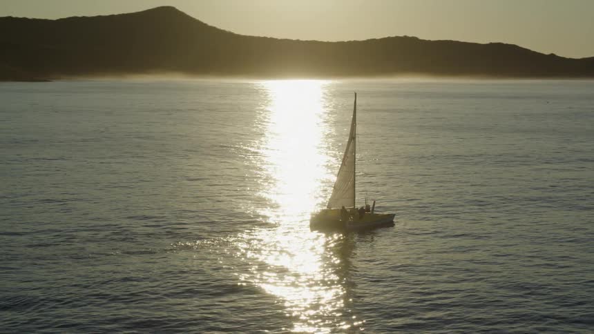 Aerial view of a small sailboat with white sails on a vast ocean at sunset