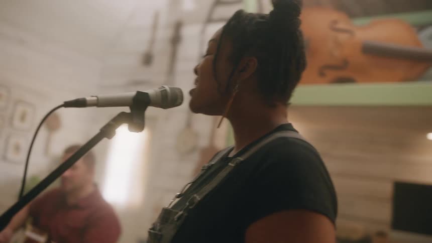 Woman in a studio singing into a microphone and next to her a man sits playing the guitar