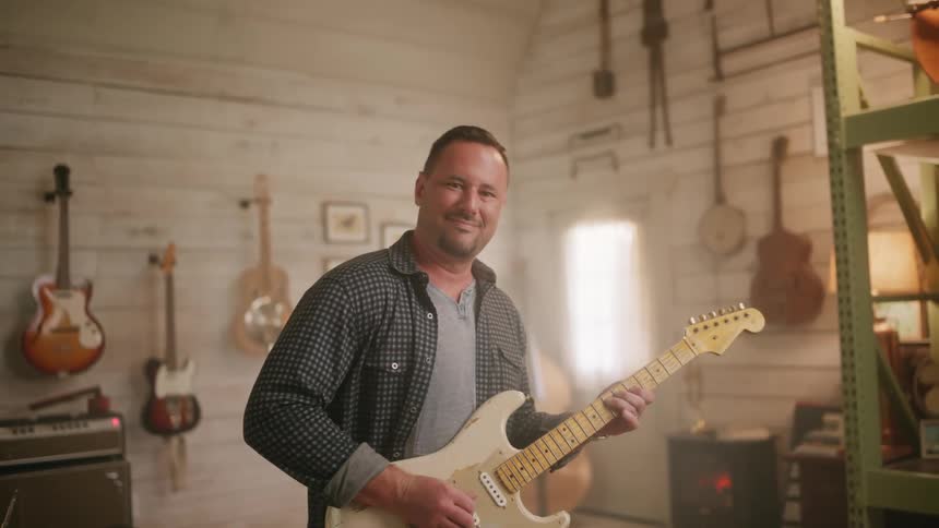A man playing a guitar surrounded by other guitars in a room