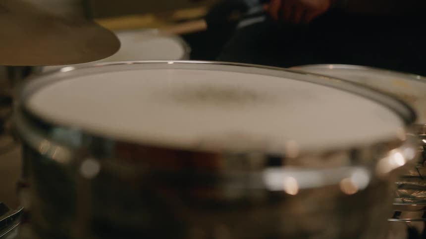 A drummer playing in a band sits at a drum set in a small studio surrounded by instruments