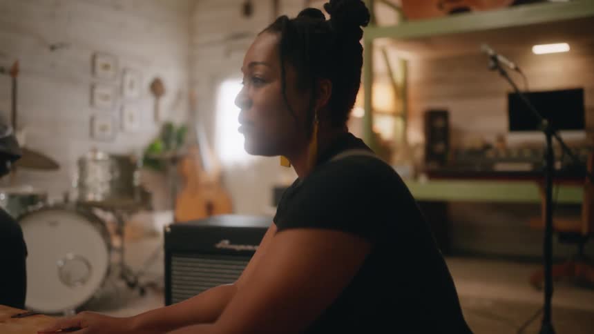 A woman hand drums on a table while singing in a room full of instruments