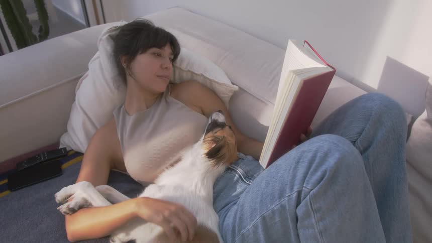 Woman lying down reading a book and petting her dog