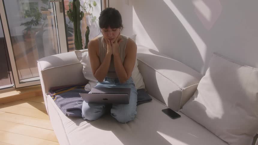 A woman is kneeling on her couch using a laptop