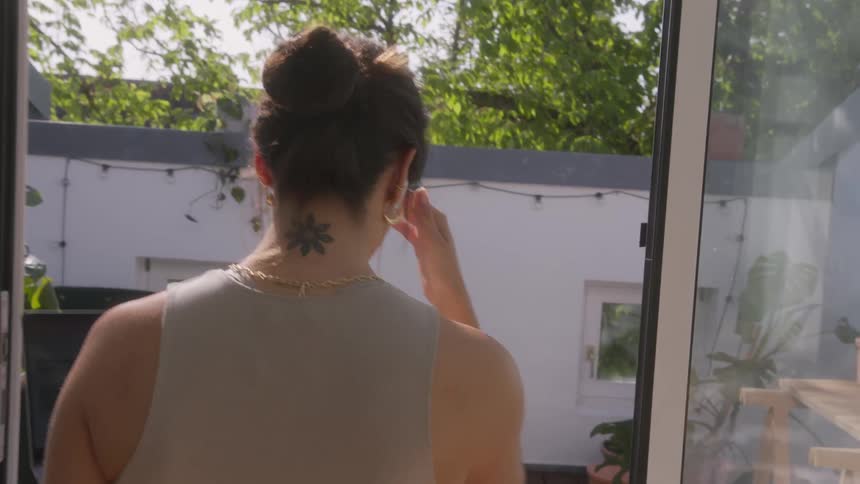 A woman in a beige shirt and jeans carries her laptop through a room while on a video call