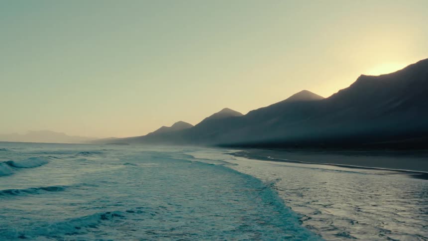 Setting sun casts the mountains into silhouettes as the surf rolls onto the beach