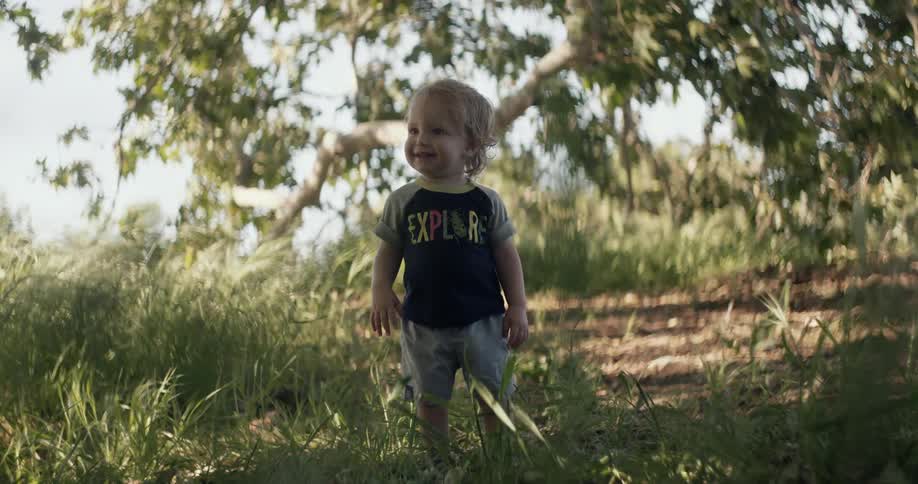 A toddler boy is standing in a wooded area on a sunny day smiling