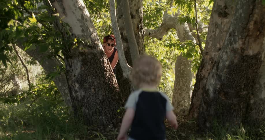 A boy is walking towards his mother who is hiding behind a tree