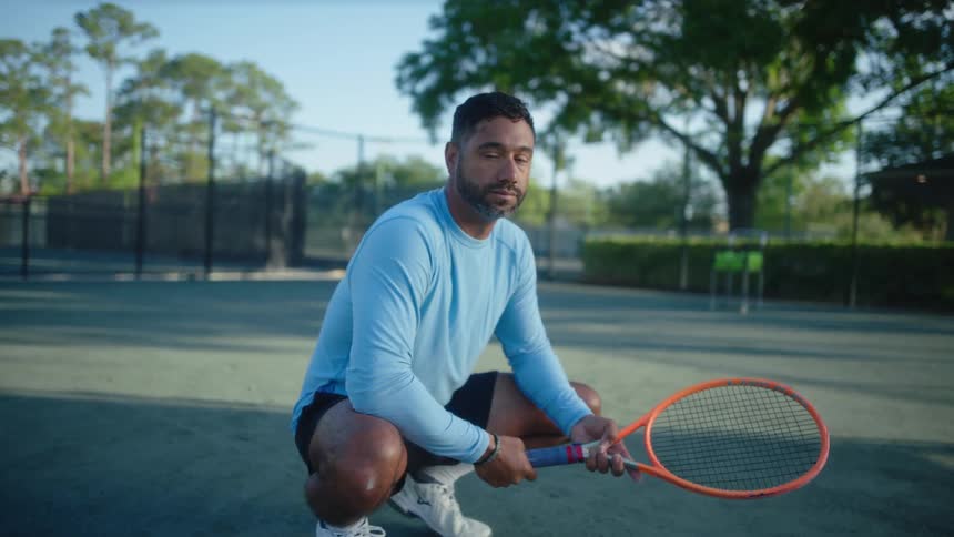 A man crouches on a tennis court and spins his racket