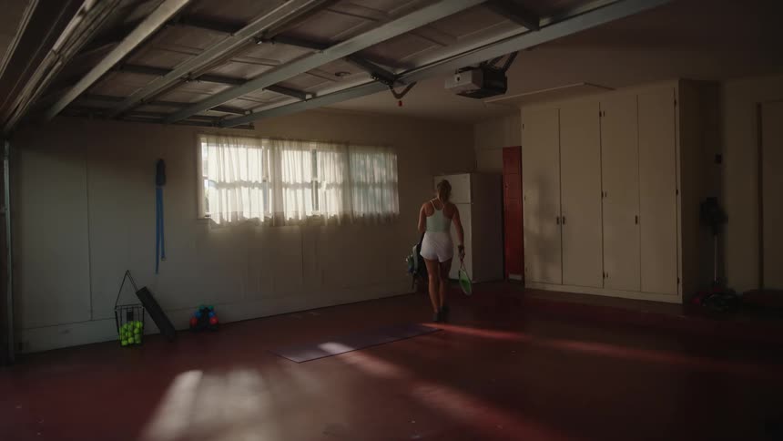 A blonde woman opens a garage door and then sits on the floor and stretches her leg