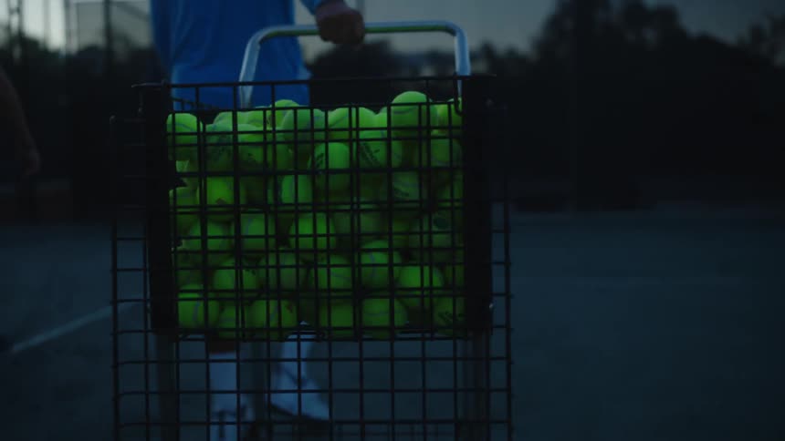 A man drags a cart full of tennis balls onto the court