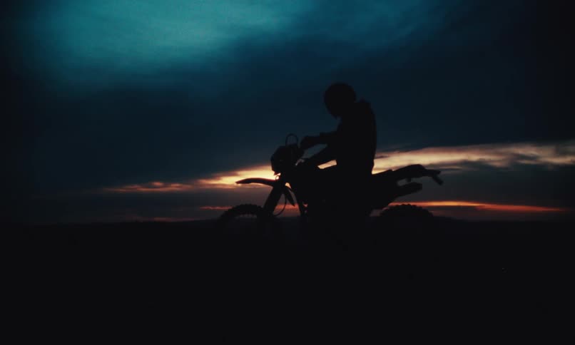 Silhouette of a man putting on his helmet and riding his motorcycle against a dark dawn sky