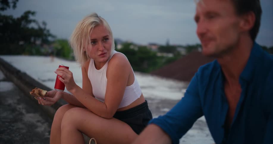 A blond man and woman are sitting and talking while the woman is eating a dessert and holding a can