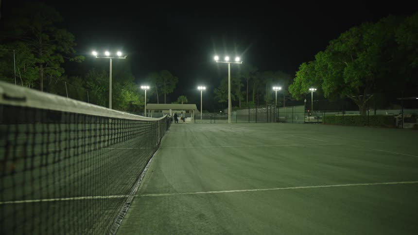 A tennis court at night is illuminated by large headlights