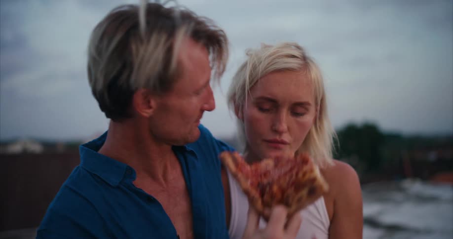 Man shares pizza with woman and kisses her while they stand hugging and talking outdoors
