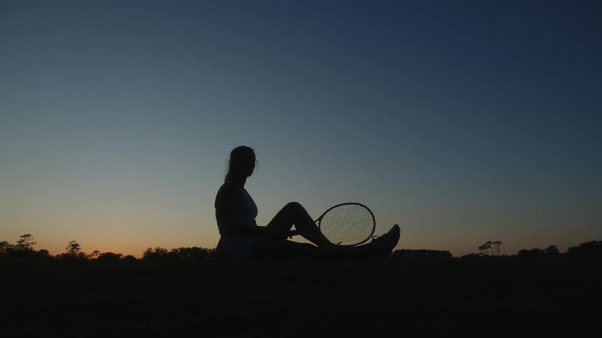 Silhouette of a seated woman holding a tennis racket against a dusk sky