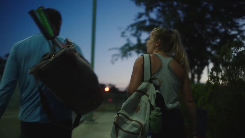 Tennis players carrying rackets walk past tennis court at night