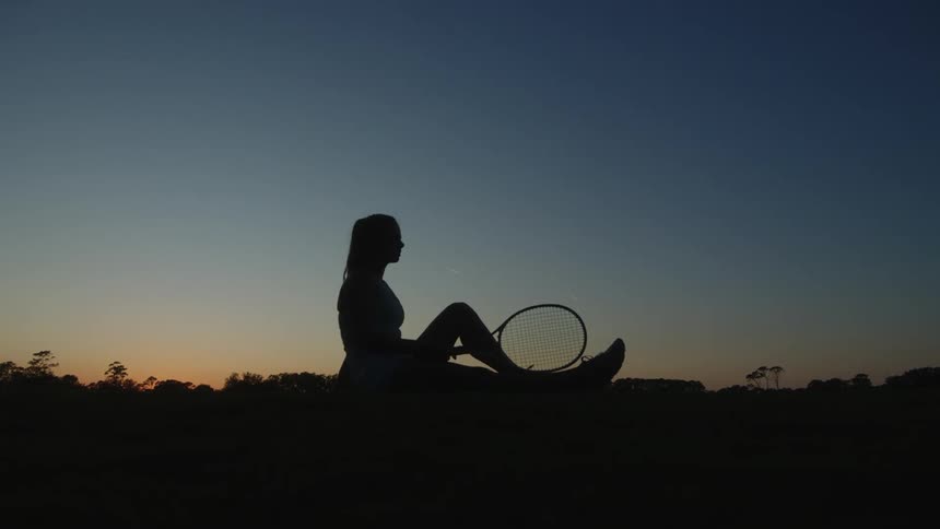 Silhouette of a seated woman holding a racket against a dusk sky