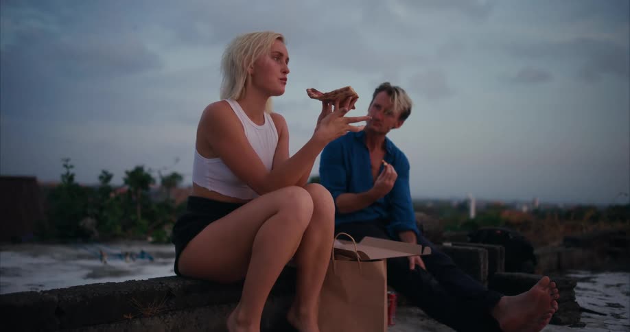 A young couple sitting on a rooftop eating pizza at evening