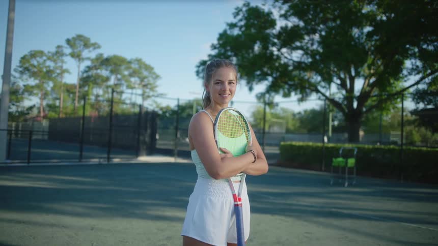 Female tennis player stands smiling while holding a tennis racket on a tennis court