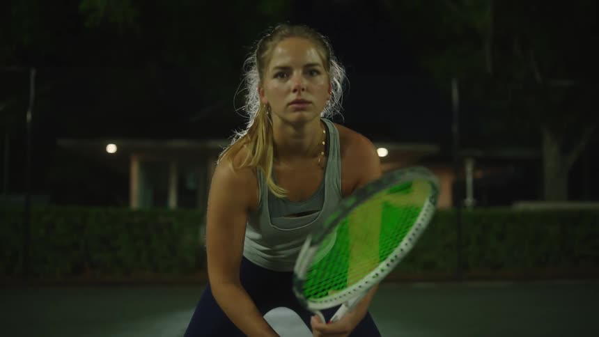 Blonde woman with concentration facial expression holding a racket on a tennis court at night