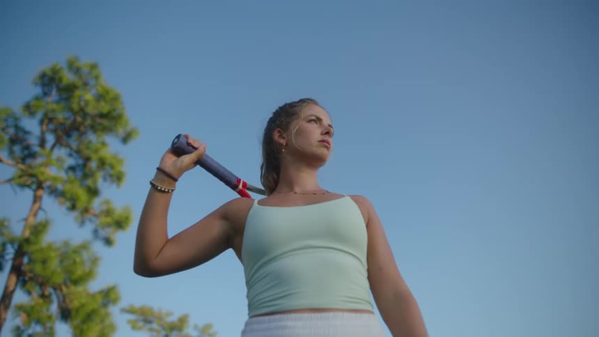 A tennis players stands outdoors holding a red racket under a clear blue sky