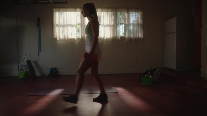 A woman holds dumbbells in each hand while doing lunges on a dimly lit room